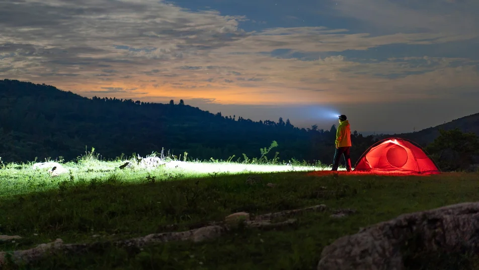 Night camper with headlamp stands beside glowing red tent in dark mountain landscape under twilight sky.