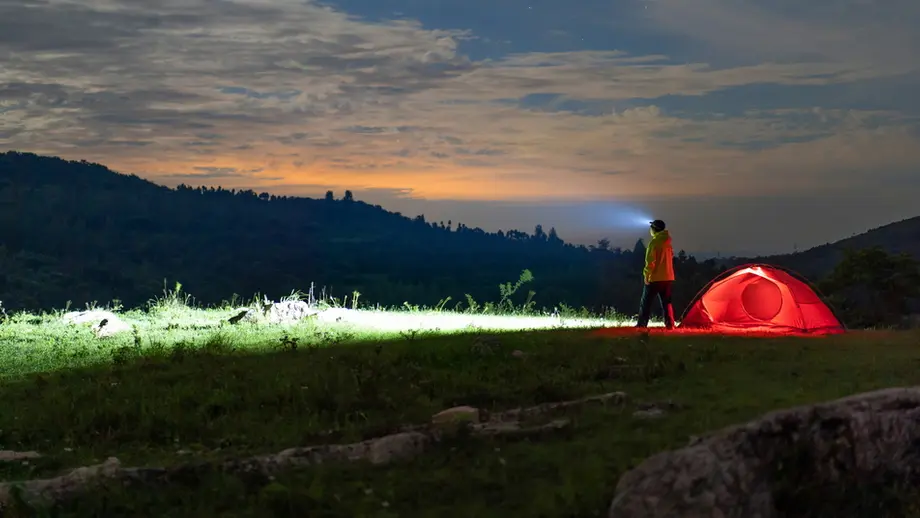 Night camper with headlamp stands beside glowing red tent in dark mountain landscape under twilight sky.