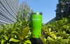 Green Olight stainless steel tumbler with red splatter pattern outdoors surrounded by green bushes under a blue sky