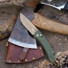 Olive green folding knife with tan blade beside a rustic axe head and brown leather sheath on wood.